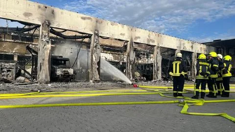 Ausgebrannte Fahrzeughalle in Stadtallendorf; Feuerwehrleute und Löschschläuche davor.